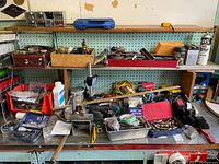 Full view of metal workbench with multiple tools, hand tools, small hardware, and accessories spread across the surface and shelves.