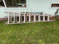 Two aluminum extension ladders laid side by side on grass against a light green building wall. Visible wear, age patina, and weather exposure evident on ladder surfaces.