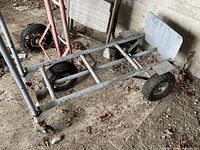 Photo showing large gray metal dolly with solid rubber wheels on a dirt floor, angled view of frame and wheels.