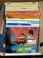 Box containing approximately 45 vintage 33 1/3 RPM records, showing a Sesame Street record prominently on top.