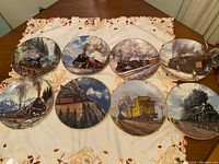 Photo showing all eight collector plates arranged on a table with a decorative cloth. Each plate features a distinct steam train scene by artist Ted Xaras.