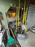 Garden tools leaning against a wall including yellow-handled shovels, a green leaf rake, and plastic snow shovel