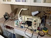 Full view of the lot items arranged on a countertop showing the sewing machine, irons, lamps and various sewing accessories scattered around.