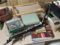 Wide view of area showing wooden stitching frame, metal green tin with sewing attachments, books, and black metal stitching clamp tool on table surface