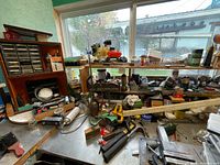 Top-down view of a cluttered workshop bench covered in multiple hand tools such as hammers, screwdrivers, and pliers, alongside small plastic and metal containers holding screws and bolts.
