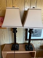 Pair of vintage lamps with black glossy turned bases and cream square lampshades on wooden table against wood-paneled wall.