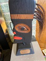 Full front view of the carved wooden Tlingit blanket ornament on stand showing face details and attached hair fibers.