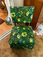 Front view of vintage low chair showing floral patterned upholstery in green, blue, and yellow tones, cushioned seat and backrest, and fabric skirt covering base.