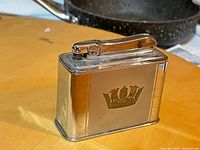 Photo of front of vintage Colibri table lighter showing crown emblem and metal finish.