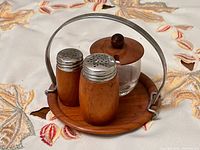 Full view of the salt and pepper set with the glass jar and teak tray with metal handle on a fabric background.