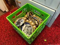 Green crate filled with stacked vintage plaques showing ornate gold rope borders and varied painted images.