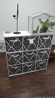Full view of white cubby storage unit with nine black and white patterned canvas bins in a 3x3 layout on wooden floor with some decor on top.