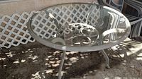 Round outdoor metal dining table with glass top and umbrella hole, placed on patio floor with lattice background.