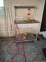 Weathered wooden potting hutch with removable lattice tops, metal tomato cages in red and green, red glass hummingbird feeder hanging, small green planter pot, and garden tools on top.