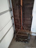 Photo of push reel lawn mower with V-shaped padded handle grips collapsed against the wall showing metal frame and blades.