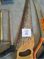 Photograph shows a collection of saws and cutting tools including a black-handled hacksaw, a large wood saw with a wooden handle, and an orange Fiskars pruning saw.