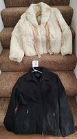 Both coats displayed on carpeted stairs: top off-white rabbit fur coat with shawl collar and tie belt, bottom black London Fog coat with zippered pockets and button closures.