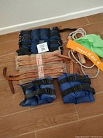 Three sets of wrist/ankle weights shown on floor, including dark blue 5 lb weights, blue 2.5 lb pair, tan leather adjustable weight straps, and two stretch bands (green and beige).