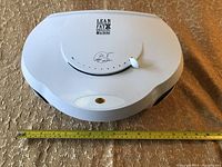 Top-down view of the white oval George Foreman grill with a measuring tape underneath showing about 14 inches in width on the carpeted floor.
