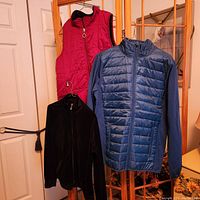 Three jackets hanging on a wooden folding screen: red sleeveless puffer vest, black zip-up jacket, blue zip-up puffer jacket with knit sleeves