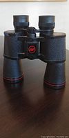 Front view of Argus binoculars showing dual eyepieces, textured black finish, red trim details, and central focus adjustment with red logo.