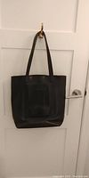 Full view of black leather tote bag hanging on a hook showing the overall shape and handles.