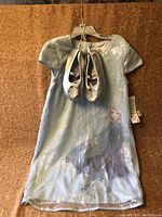 Full front view of the Elsa dress with the silver shoes hanging on hanger in front of a brown backdrop.