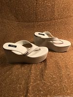 Side view of white DKNY platform sandals on a brown textured surface, showing the overall design and height of the platforms.