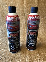 Two cylindrical spray cans with black caps labeled FW1 FastWax Cleaning Wax, red car image, and usage directions.