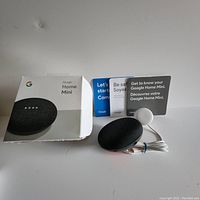 Google Home Mini smart speaker in black with original box, power adapter, and instruction booklets.