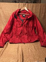 Front view of red Helly Hansen jacket on hanger against beige backdrop.