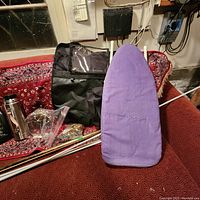 Photo showing small purple-covered ironing board, vacuum thermos, and black vehicle organizer on red patterned surface