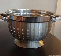 Front side view of the shiny large stainless steel colander showing perforations, handles, and base.