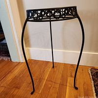 Full view of vintage black metal plant stand with round frame and three curved legs on hardwood floor.