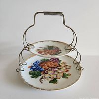 Front view of the two-tier cake plate showing painted fruit on two porcelain plates with metal stand and spring handle.
