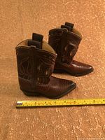Side angle view of two brown miniature cowboy boots placed on carpet with measuring tape showing approximate length.