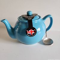 Blue stoneware teapot with matching lid and attached label, and a detailed metal spoon beside it.