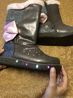Side view of metallic silver Skechers girl's winter boots showing knit cuff, pink bow, glitter heel, and lights on sole illuminated in different colors.