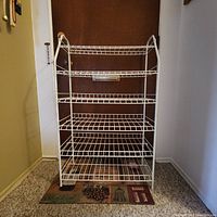 Front view of the 6-tier white metal wire shelving unit standing on a mat against a closed door inside a room.