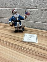 Front view of the 2003 Lenox Santa figurine standing on a wooden surface, holding an American flag and toy with informational card placed in front.
