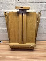 Front view of folded wooden art easel with side panels collapsed and canvas holder clamp at top center.