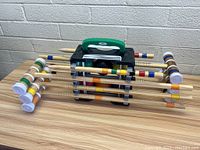 View of eight wooden croquet mallets with colored bands, arranged on a black plastic stand with green handle against a brick wall background