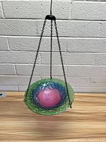 Hanging glass bird bath with a multicolored, textured glass bowl suspended by three black metal chains connected to a top hanger, against a white brick wall.