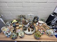 Large collection of assorted snow globes and home decor items displayed on a wooden table against a beige brick wall. Items include various snow globes, figurines, and decorative pieces like a teapot and fruit display.