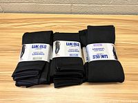 Six pairs of black Lukees fleece-lined leggings folded with original paper bands showing brand and product details, arranged on wood surface.