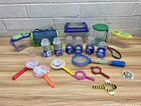 Lot of various bug catching toys displayed on wooden surface including containers, nets, magnifiers, and carry bag