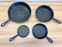 Lot showing six cast iron pans of various shapes and sizes arranged on wooden surface, mostly round with handles.