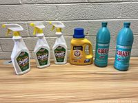Six cleaning products displayed against a beige brick wall: three white spray bottles of Fabuloso All Purpose Cleaner with Bleach, one yellow bottle of Arm & Hammer detergent, and two teal bottles of Cloralen bleach.