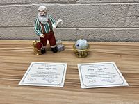 Porcelain Santa figurine standing on a wood surface against a brick wall holding a scroll labeled 'Santa's Flight Plan', next to a dog figurine and a gift box, along with a decorative globe topped with stars and bells, and two Lenox certificates in front.