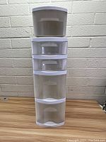 Front view of white plastic storage shelf with five drawers on a wooden surface in front of a brick wall.